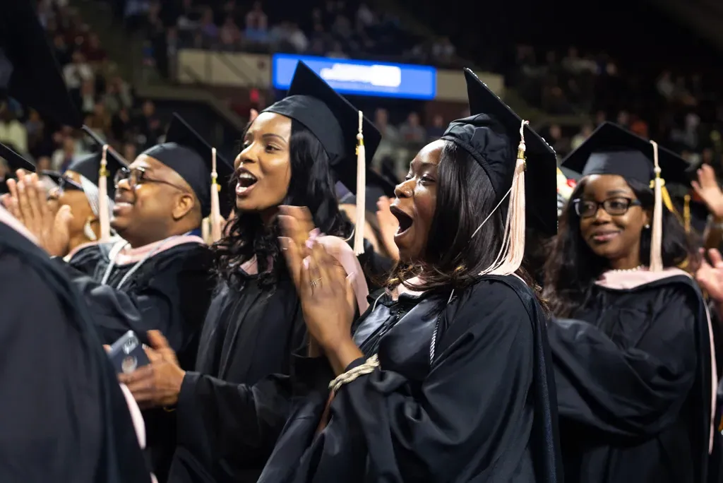 a group of U N E student wearing graduation regalia stand and applaud